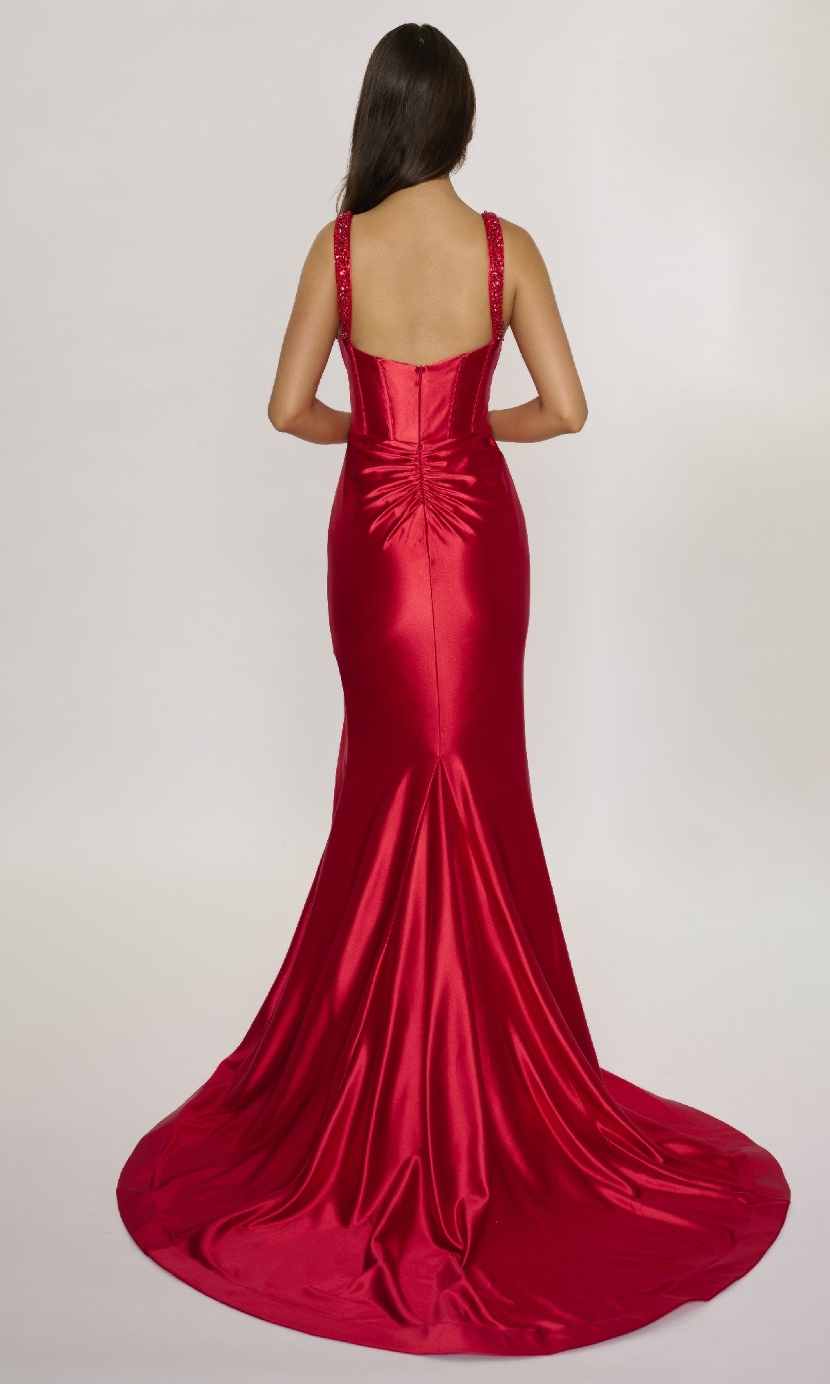 A woman with long dark hair faces away, wearing the Nina Canacci 3271 long formal dress in red satin, featuring a fitted corset bodice, dramatic train, and ruched detailing at the back.